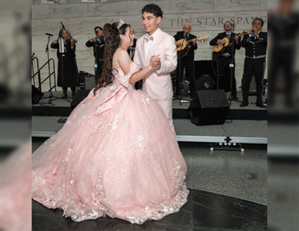 Photograph of a young girl in a pink gown and sequined tiara dancing with a boy in a tuxedo while a mariachi band plays in the background.
