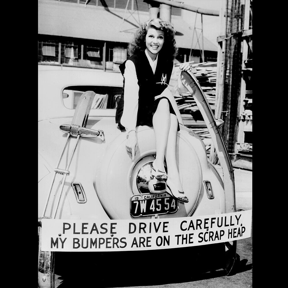 Rita Hayworth sitting on the back of a car with a sign under her feet displaying the text: Please drive carefully my bumpers are on the scarp heap.