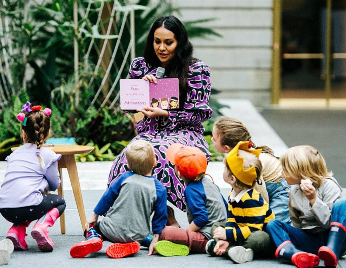 Storybook author in black and purple dress reads to a group of children seated around her on the floor.