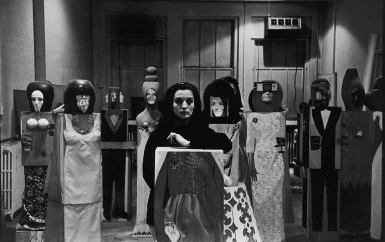 A dramatically lit black & white photograph of Marisol Escobar wearing a black top standing among several of her life-size, painted and carved wooden sculptures.